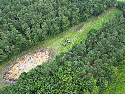 Luftbild, Schrägaufnahme: v. o. Wald, große runde Mulde inmitten von Grünland, Wald. 