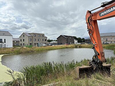 Ein Bagger steht am Teich, dahinter stehen Häuser.
