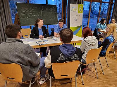An einem Tisch vor einer Schultafel geben Margarita Keller und Lennart Fietz den drei vor dem Tisch sitzenden Schülern Infos zum Wasserverband.