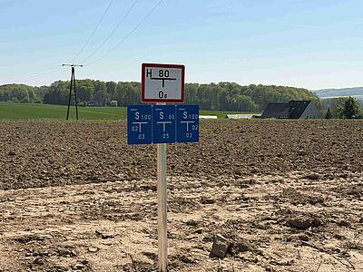 Schilder vor Ackerfläche: oben weißes Hydrantenschild, darunter blaue Schilder, die Angaben zum Schieber machen