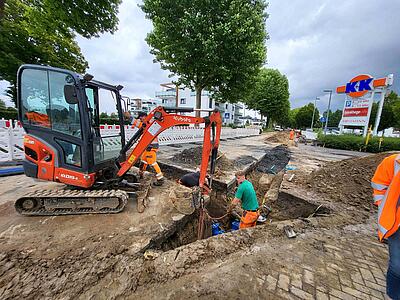 Baggerarbeiten an einer Straße. Ein Bauarbeiter steht in der Baugrube und platziert eine Leitungsarmatur.