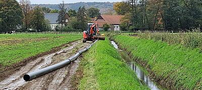 Eine Druckrohrleitung wird auf einer Grünlandfläche verlegt. Im Hintergrund ein Bagger.
