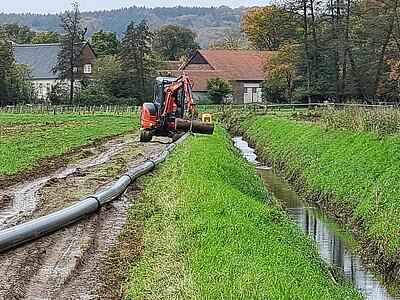 Erdarbeiten zur Verlegung der Leitung. Links Acker, danach Aushub mit Bagger und liegende Leitung, Graben, Feld. Im Hintergrund Häuser.