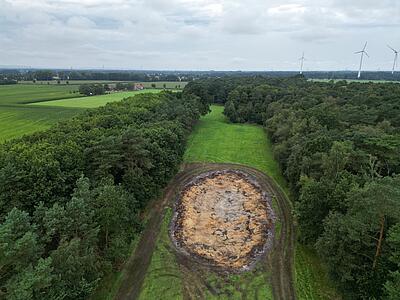 Luftbild: v. l. Grünland, Wald, große runde Mulde inmitten von Grünland, Wald. Im Hintergrund verstreute Höfe und Windräder.